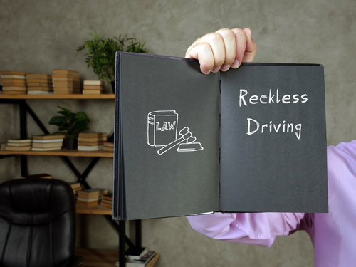 A person in a formal shirt holding an open black notebook displaying a chalk-style drawing of a law book and gavel on one page, and the words “Reckless Driving” written on the other page, in a professional office setting with bookshelves in the background.