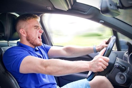 A young man in a blue polo shirt screaming while aggressively gripping the steering wheel of a moving vehicle, captured mid-action to depict reckless or emotional driving.