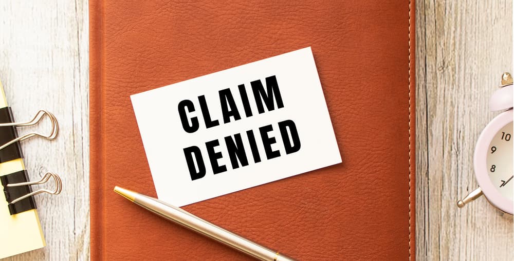 A business card with 'CLAIM DENIED' on a wooden table alongside a brown diary and pen. Business concept.
