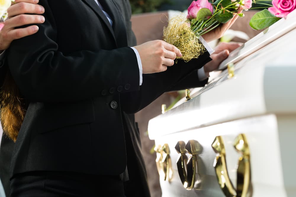 A man and woman mourn at a funeral, standing beside a casket adorned with a pink rose.