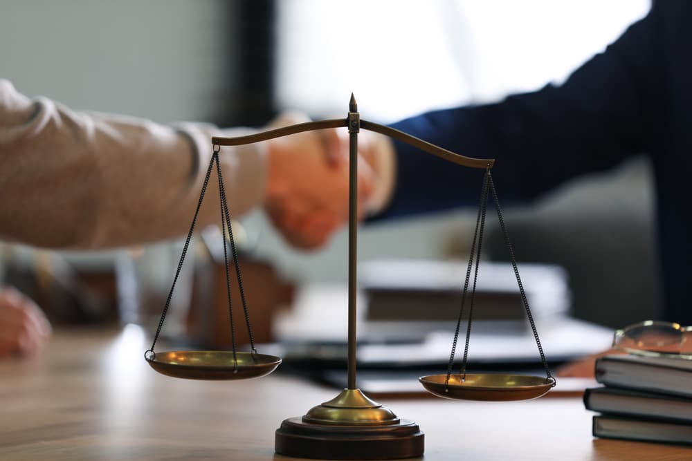 A male lawyer shaking hands with a client in an office, with the scales of justice in focus.