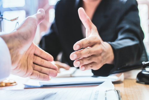 A close-up of two people extending hands for a handshake across a desk, suggesting agreement or the conclusion of a legal or professional meeting.