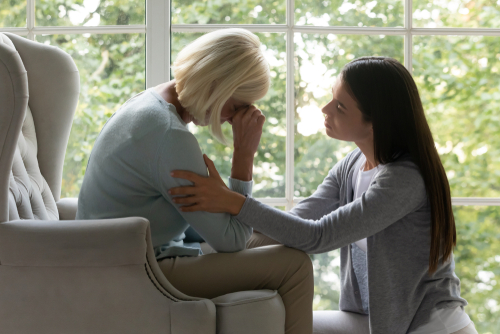 A young woman comforts an older woman who is sitting in a chair and appears to be crying. They are indoors near a window with greenery outside.