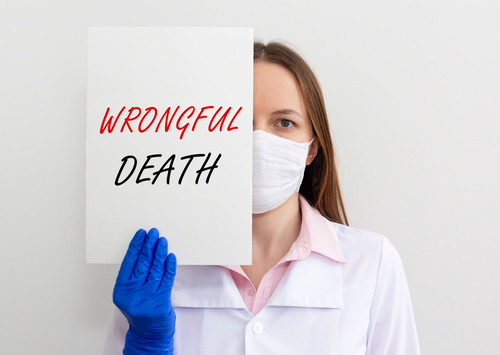 A healthcare professional wearing a mask and blue gloves holds a sign that says "WRONGFUL DEATH," with half their face visible beside the sign.