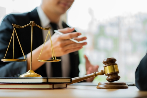 A lawyer in a suit sits at a desk with legal documents, a wooden gavel, and brass balance scales symbolising justice.