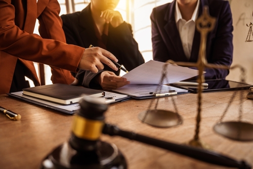 A group of professionals in business attire discussing legal documents at a table, with legal scales and a gavel in the foreground, indicating a legal consultation or courtroom preparation.