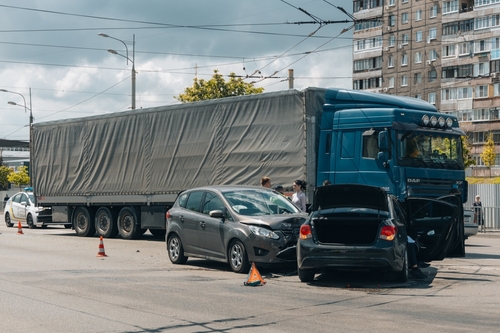 A large blue semi-truck involved in a multi-vehicle collision with two smaller cars at an urban intersection. The vehicles appear heavily damaged, and the scene is marked with warning cones, suggesting an ongoing accident investigation.