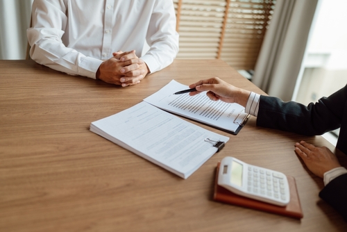 Person in a business suit pointing to a legal document during a meeting with another individual at a desk.