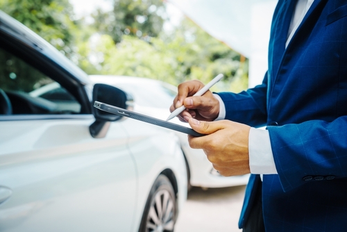 Man in a blue suit writing on a clipboard next to a white car.