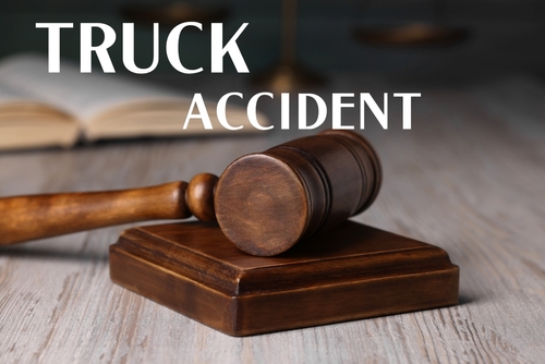 A close-up of a wooden judge’s gavel resting on a sound block, with the words “TRUCK ACCIDENT” written in bold white text above. The background includes a blurred open law book, symbolizing legal proceedings related to a truck accident.