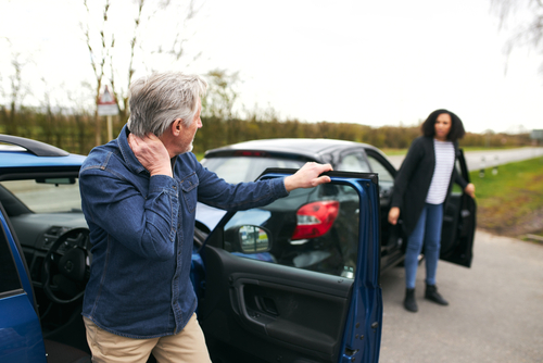 An older man holding his neck in pain after a car accident, standing next to his open car door while another driver approaches from the background.