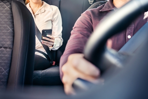 Close-up view of a female rideshare passenger in the back seat using a smartphone while the male driver focuses on the road.