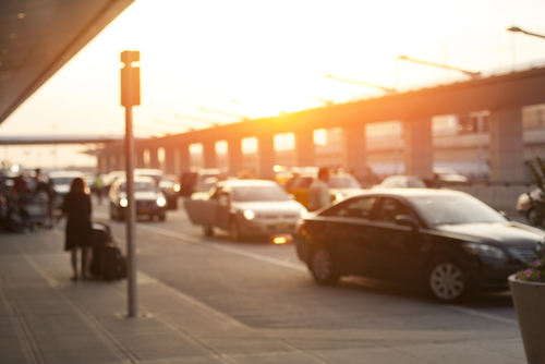Blurred image of cars lining up at an airport pickup zone during sunset with people walking on the sidewalk.