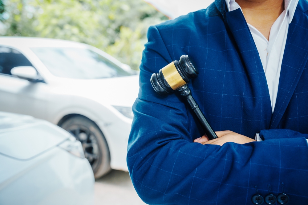 Lawyer in a blue suit holding a gavel across his chest while standing near parked cars, symbolizing justice and legal authority.