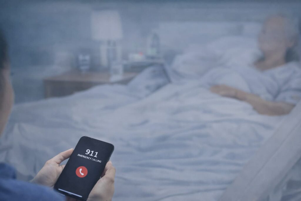 Person dialing 911 on smartphone in nursing home room during suspected medical emergency involving elderly resident.