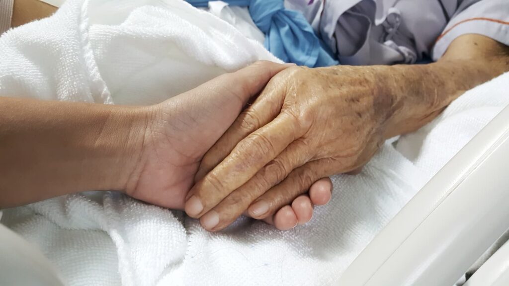 Family member holding elderly nursing home resident&rsquo;s hand in hospital bed, symbolizing concern about sepsis and bed sore neglect.